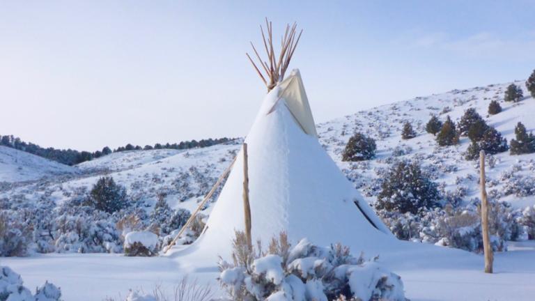 A snow-covered tipi sits in a hilly landscape dotted with trees and bushes against a blue sky. Partially obscured.