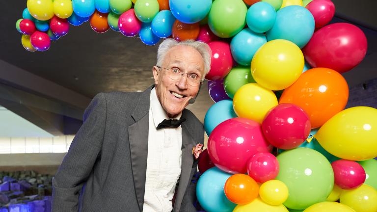 A performer wearing a tuxedo stands in front of a display of multi-coloured balloons. Partially obscured.