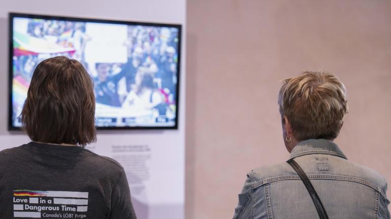 Two people stand with their backs to us and watch a video. The wall holding the video monitor features rainbow colours.