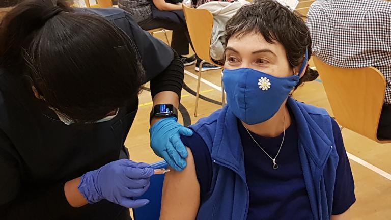 A healthcare worker prepares to give a vaccine to a patient. Both people are wearing medical face masks. Partially obscured.