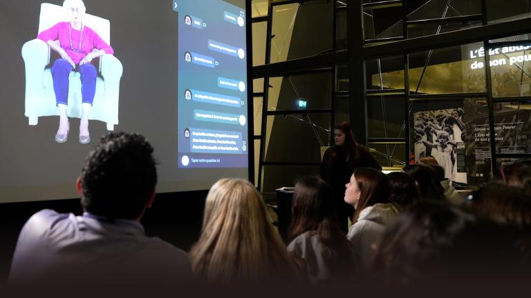 Audience members view a large projection screen displaying an elderly woman in a purple top and blue pants sitting in a white armchair, alongside an interactive message interface. The presentation takes place in a modern space with geometric glass architecture, and a presenter stands to the right of the screen. Partially obscured.