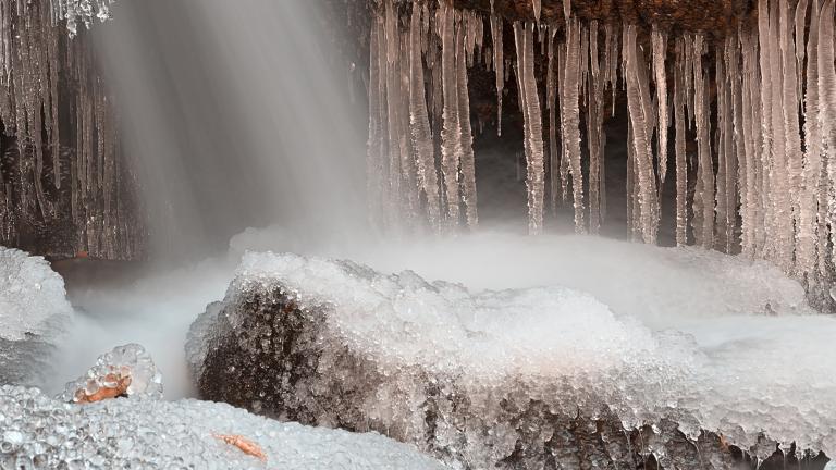 Water sprays onto rock formations from an unseen opening above, causing a dramatic array of icicles and mounds of ice on boulders. Partially obscured.