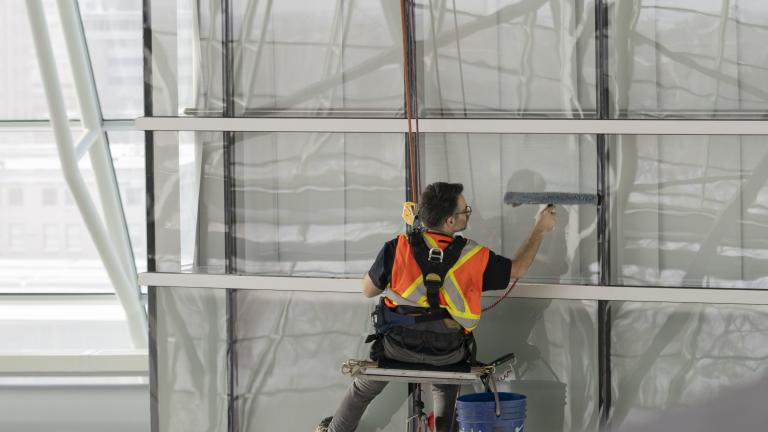 Male worker wearing safety vest suspended from ropes cleaning windows from a high height. Partially obscured.