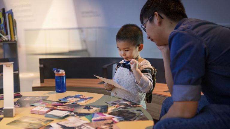 A young child interacting with photos on a table. An adult sits beside him. Partially obscured.