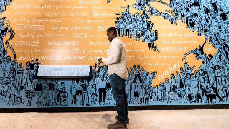 A man stands in front of an illustrated blue and orange a map of Canada. Partially obscured.