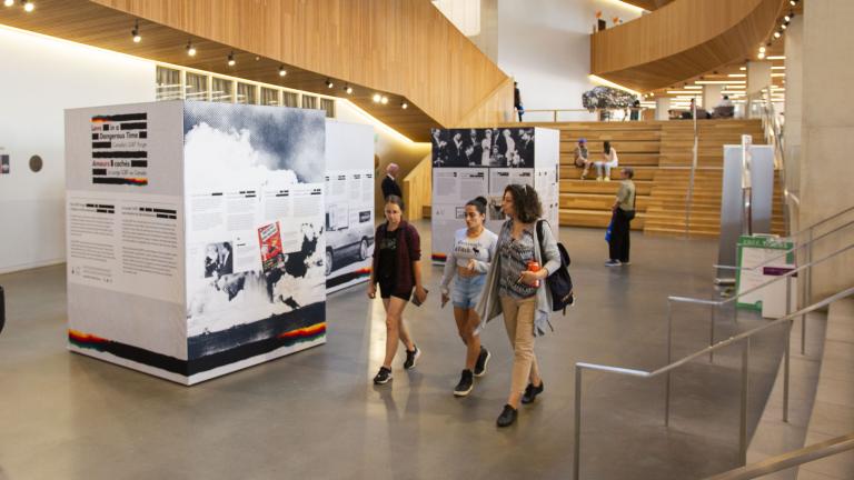 Interior view of a white atrium with wooden levels above. Visitors are walking past three standalone informational displays. The displays include text and images set against a backdrop of wooden architecture and a staircase. Some visitors are engaged in conversation, while others interact with the exhibition.