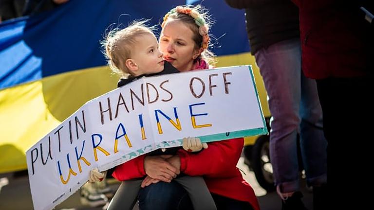 A child is holding a protest sign that says: “Putin, hands off Ukraine.” The child is sitting on their caregiver’s lap. Behind them is a Ukrainian flag. Partially obscured.