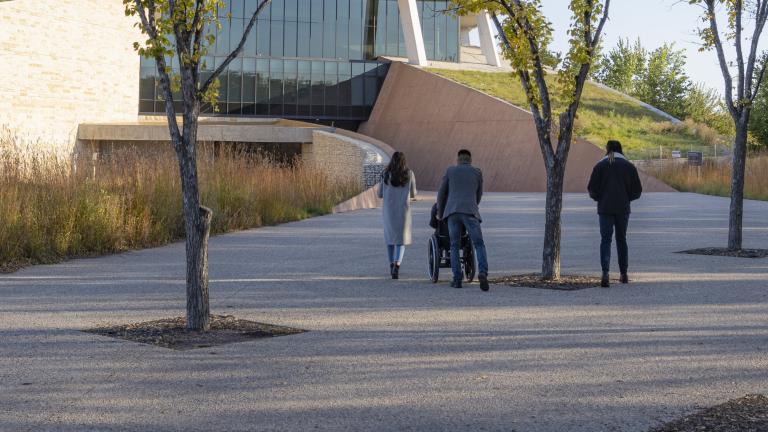Four individuals follow the path toward the Canadian Museum for Human Rights on a fall day. Three are on foot and one is in a wheelchair. Partially obscured.
