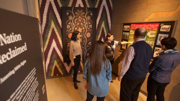 A guide speaks to a small group of people in a museum exhibit space. The space includes a wall featuring a Métis artwork and a text panel that reads “A Nation Reclaimed”. Partially obscured.