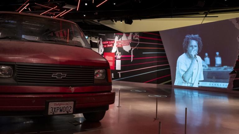 Red Chevrolet van in an exhibition space with video by Rage Against the Machine playing in the background. Partially obscured.
