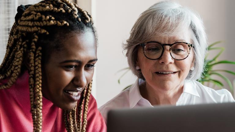 A young woman who looks to be in her teen years smiles as she works at a computer alongside an older, white-haired woman who is also smiling. Partially obscured.