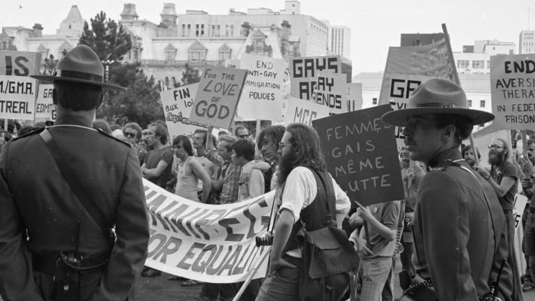 A large group of protestors march through a city while two uniformed RCMP officers keep watch. In this black-and-white image, the protestors' signs and banners call for an end to discriminatory practices against members of the 2SLGBTQI+ community. Partially obscured.