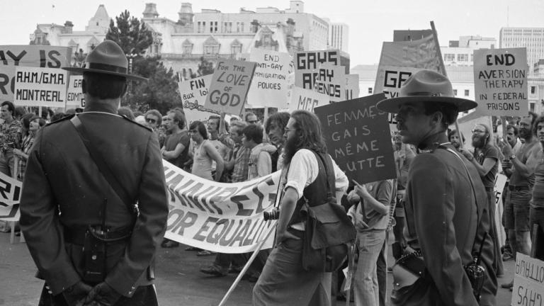 A large group of protestors march through a city while two uniformed RCMP officers keep watch. In this black-and-white image, the protestors' signs and banners call for an end to discriminatory practices against members of the 2SLGBTQI+ community. Partially obscured.