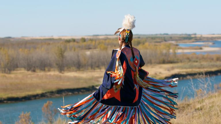 A Indigenous dancer in brightly coloured Fancy Shawl regalia beside a river. Partially obscured.