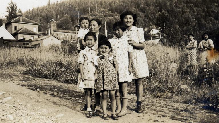 Black-and-white image of women and children standing on a gravel road in front of a field. Partially obscured.