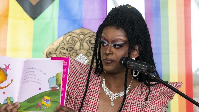 A drag queen in heavy makeup and a wig of long tiny braids holds up a children’s book to show an audience. The backdrop behind her is a pastel rainbow. Partially obscured.