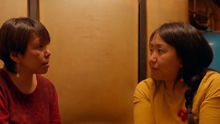 Two Inuit women sit in a dim room facing one another in intimate conversation. One is wearing a red sweater, the other a yellow sweater. Partially obscured.