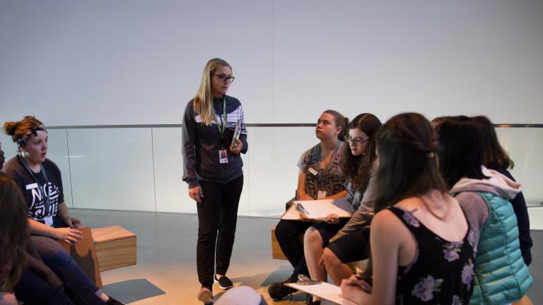 Students sit on benches in a circle with clipboards on their laps. A woman is standing and talking to them. Partially obscured.