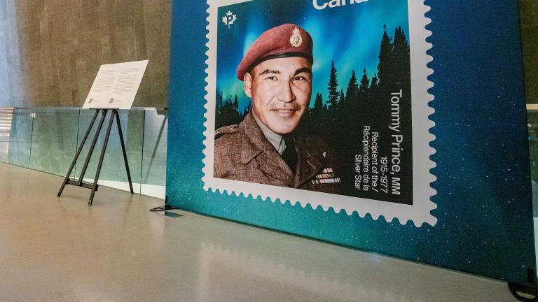 An enlarged Canadian stamp showcases a man with a red military beret with an aurora borealis and darkened forest in the background. Partially obscured.