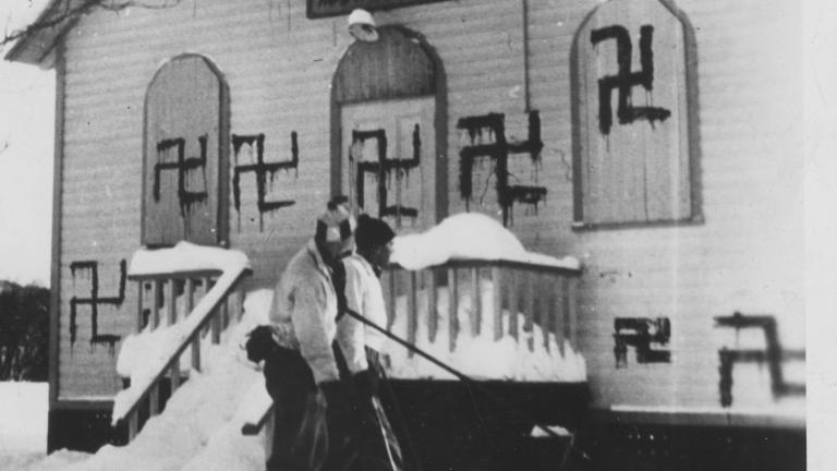 Two people play hockey in front of a synagogue with several swastikas painted on it. Partially obscured.