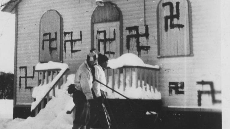 Two people play hockey in front of a synagogue with several swastikas painted on it. Partially obscured.
