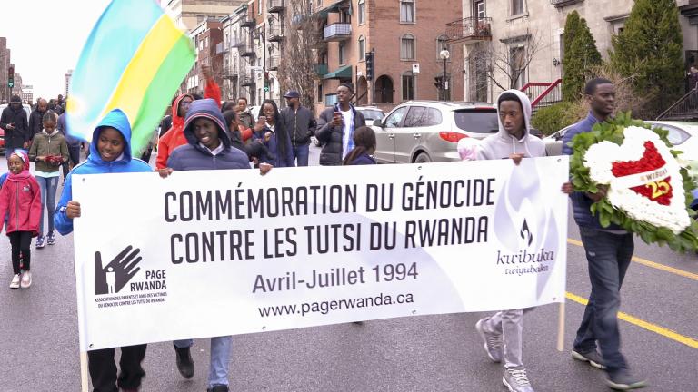 A group of people walking down a street with sign that reads “Commémoration du génocide contre les Tutsi du Rwanda. Avril-Juillet 1994. www.pagerwanda.ca”. Partially obscured.