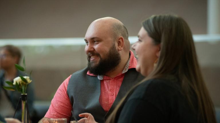 A man and woman sit together at a dinner table, smiling warmly as they listen to other guests. The man wears a salmon shirt with a gray vest, and there are small candles and drinks on the table, creating a cozy intimate atmosphere. Partially obscured.