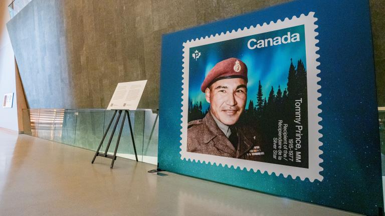 Un grand affichage d'un timbre représentant le sergent Tommy Prince est visible sur le côté droit de l'image. Une passerelle située devant le panneau d'affichage mène à gauche de l'image, dans la galerie Perspectives indigènes. Partially obscured.