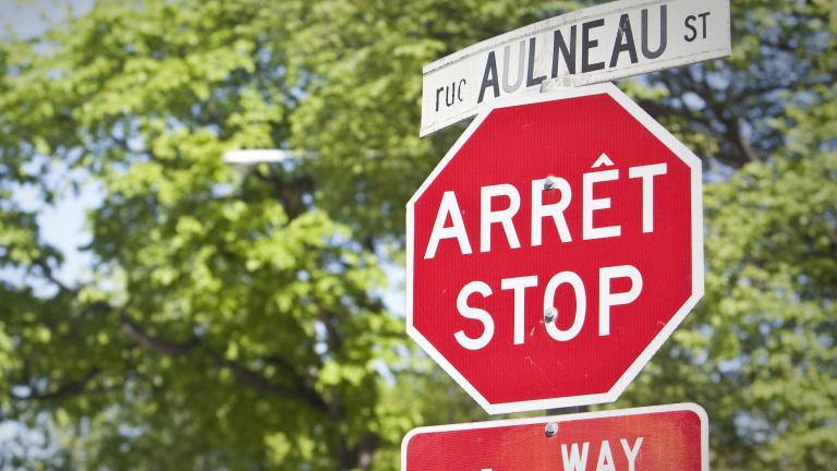 A stop sign and street sign in both English and French are seen in front of trees. Partially obscured.