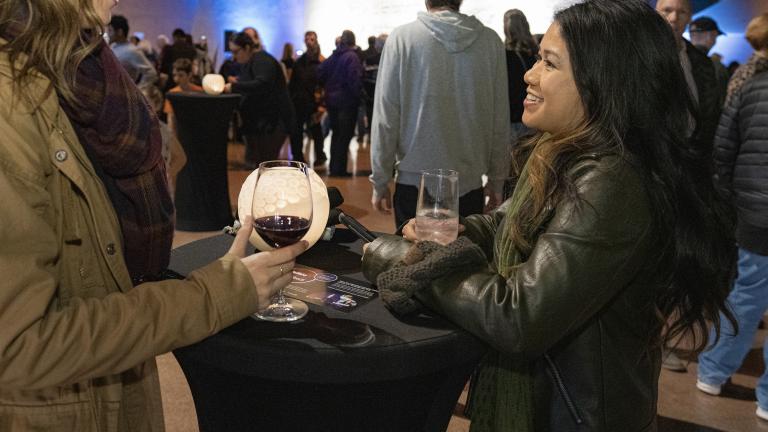 Two smiling women drink wine in a large room full of people. Partially obscured.