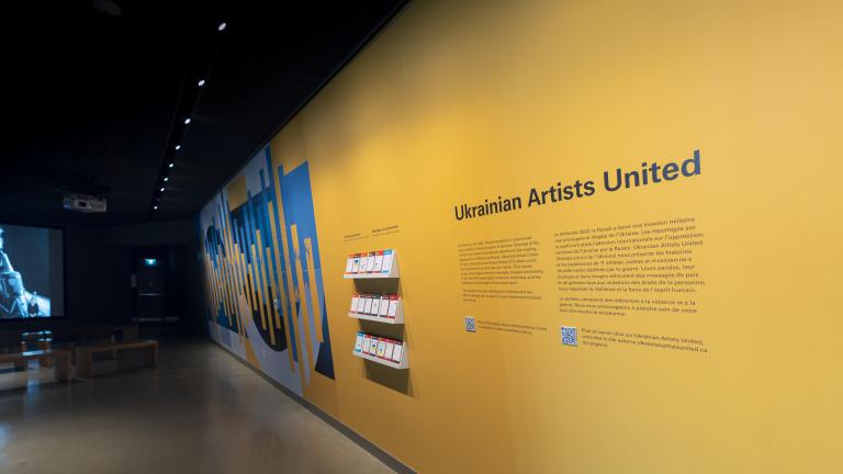 A yellow wall with the words "Ukrainian Artists United" and a dynamic blue and yellow graphic is right side of the image. On the left side, an image is projected on a screen showing a person with headphones and an open mouth, as though they are singing. Benches for sitting are placed in front of the screen. Partially obscured.