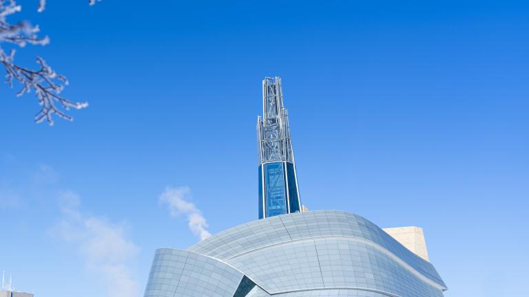 An outdoor winter scene with an exterior view of the Canadian Museum for Human Rights, other buildings in the distance and trees covered in frost. Partially obscured.