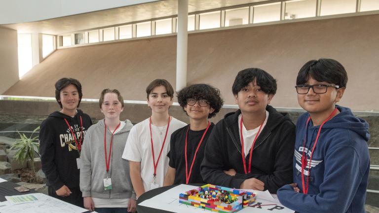 Six students in a museum gallery are standing behind tables on which are placed posters of their presentations. Partially obscured.