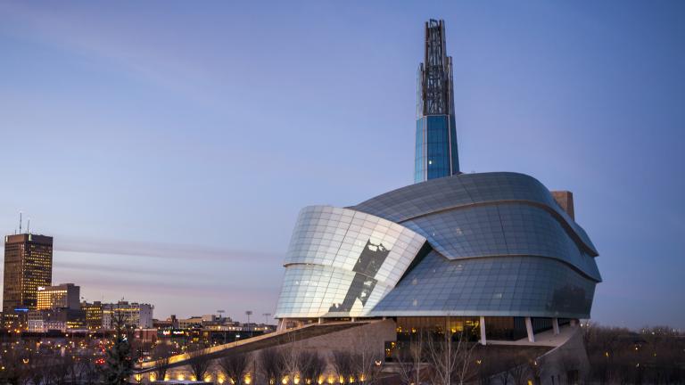 The exterior of the Canadian Museum for Human Rights, seen in the sunset against the city skyline. Partially obscured.