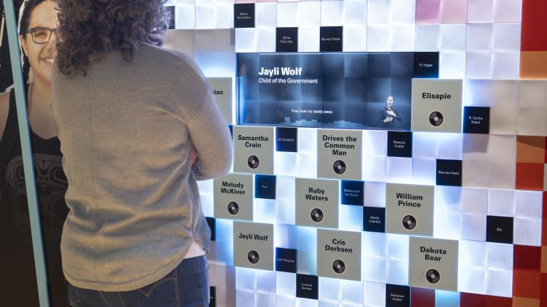 A visitor engages with an interactive wall component made up of small squares with a blue screen in the centre. Light-coloured squares peppered with black ones and grey ones containing text form a circular shape at the centre while squares in shades of red are on the periphery. To the left of the visitor is a graphic panel showing smiling faces.