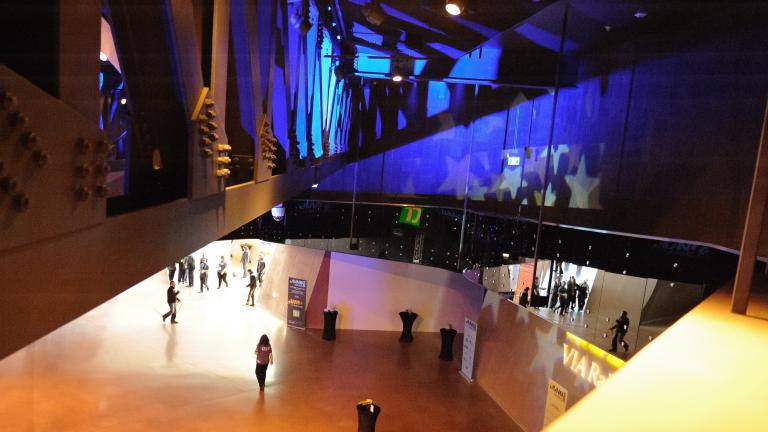 Several people stroll through a great open hall. They are viewed from a ramp above. The ramp’s metal structure and rivets are visible on the left, while on the right a gleaming mirrored wall reflects the people below plus vivid indigo lighting and white stars. Partially obscured.