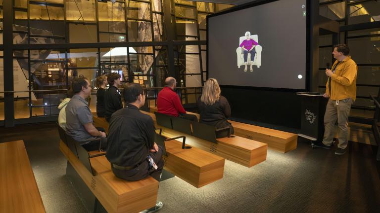 A small group of people sitting on wooden benches in a theatre face a screen showing an older woman seated in a chair. A man in a yellow shirt is standing to the right of the screen holding a microphone. Partially obscured.