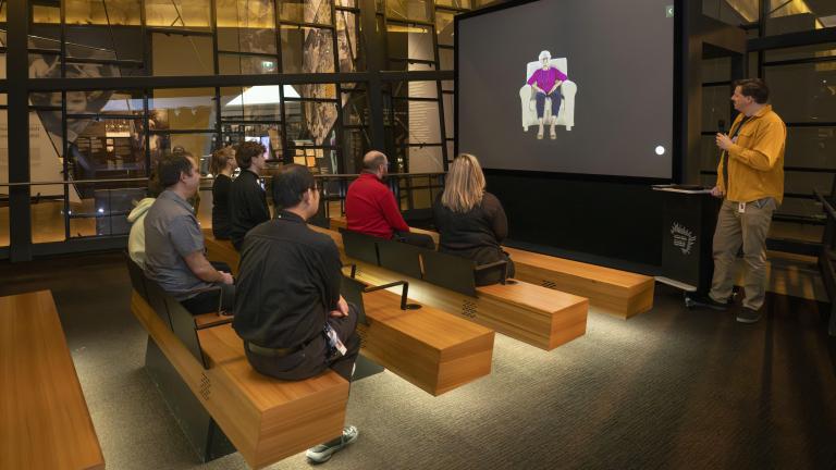 A small group of people sitting on wooden benches in a theatre face a screen showing an older woman seated in a chair. A man in a yellow shirt is standing to the right of the screen holding a microphone. Partially obscured.