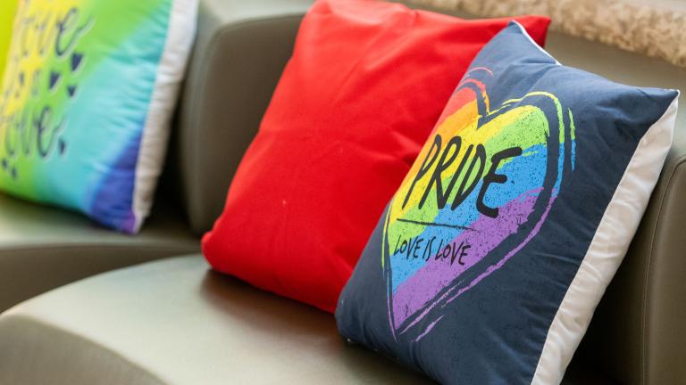 Four colourful cushions on a bench with Pride written on the first one. Partially obscured.