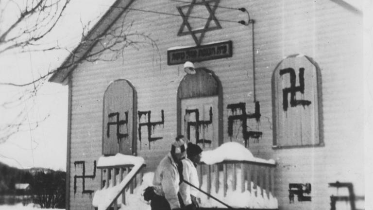 Two people play hockey in front of a synagogue with several swastikas painted on it. Partially obscured.