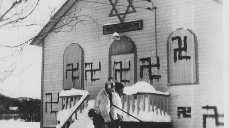 Two people play hockey in front of a synagogue with several swastikas painted on it. Partially obscured.
