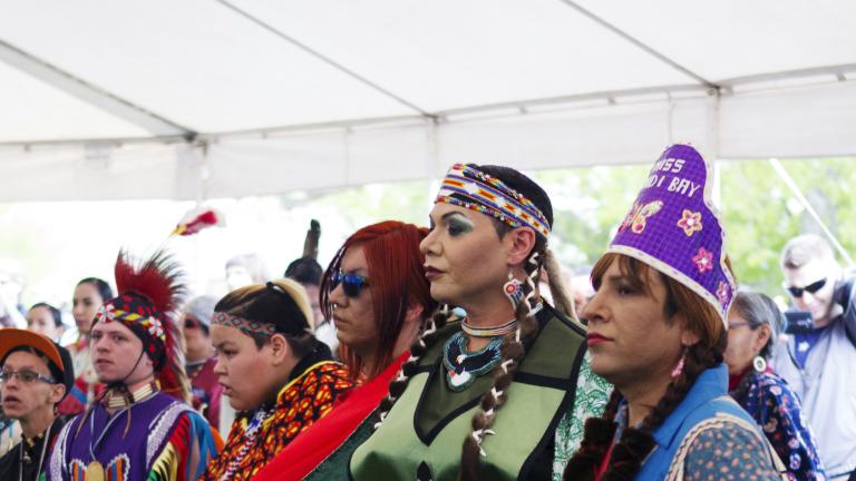 A group of people wearing colourful Indigenous regalia dancing in a line inside a tent on grass.