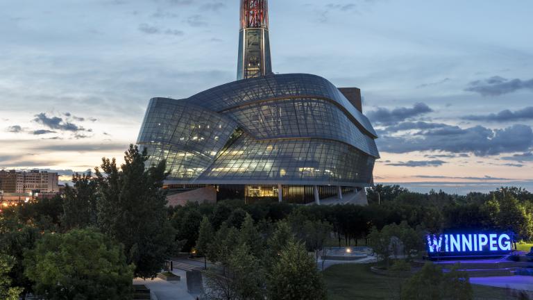 Exterior photo of the Canadian Museum for Human Rights’ glass windows and tower, taken against a prairie sky at dusk. The Winnipeg sign at the Forks is situated on the right side of the image. Multiple office towers and downtown lights are on the left-hand side of image. Partially obscured.