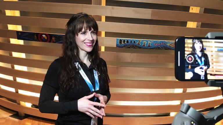 A smiling woman with light skin is filmed on a cell phone. She is wearing a lanyard with the words “Be inspired” and standing in front of an exhibit element made of horizontal strips of wood, some decorated with Indigenous art.