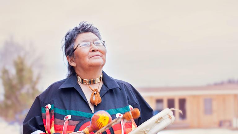 Elder Ma-Nee Chacaby stands outside surrounded by trees and cabin looking up at the sky playing a drum. 