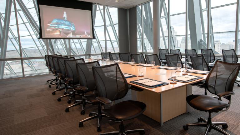 A carpeted meeting room surrounded by glass walls with a long boardroom table, chairs and a presentation screen.