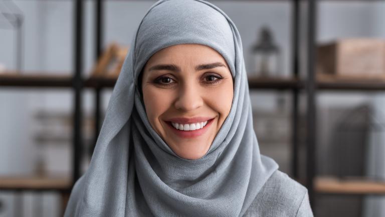 A woman wearing a blue hijab smiles toward the camera, with a bookshelf in the background.