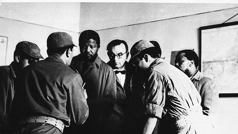 A black-and-white image of Nelson Mandela standing in front of a desk. He is surrounded by a group of five men, some of whom are wearing military clothing. One man, with his back to the viewer, appears to be speaking to Mandela, while the other men are looking at something on the desk.