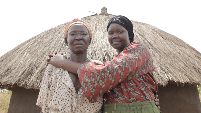 Two women are standing in front of a hut with a straw roof, looking at the camera. The younger woman on the right is smiling and embracing the one on the left.