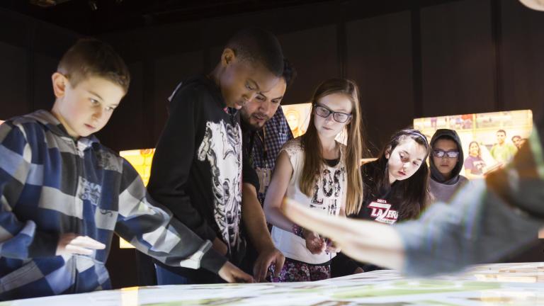 A group of students look at the top of an interactive table emitting white light.